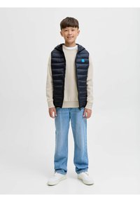 Chaleco acolchado negro con capucha, con una etiqueta azul, sobre un suéter crema. Jeans rectos azul claro y zapatillas blancas completan el atuendo.