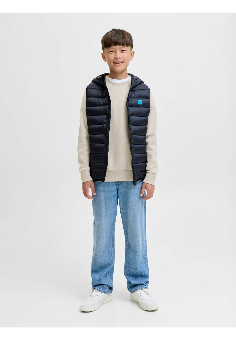 Chaleco acolchado negro con capucha, con una etiqueta azul, sobre un suéter crema. Jeans rectos azul claro y zapatillas blancas completan el atuendo.