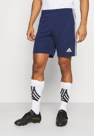 Mężczyzna ubrany w granatowe spodenki Adidas, białe sportowe skarpetki z czarnymi wzorami oraz czarne korki piłkarskie Adidas Nemesis, stojący na szarej podłodze.