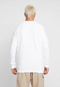 Sudadera blanca de manga larga, con puños y dobladillo acanalados. Textura suave, diseño sencillo y corte holgado, mostrada desde la parte posterior.