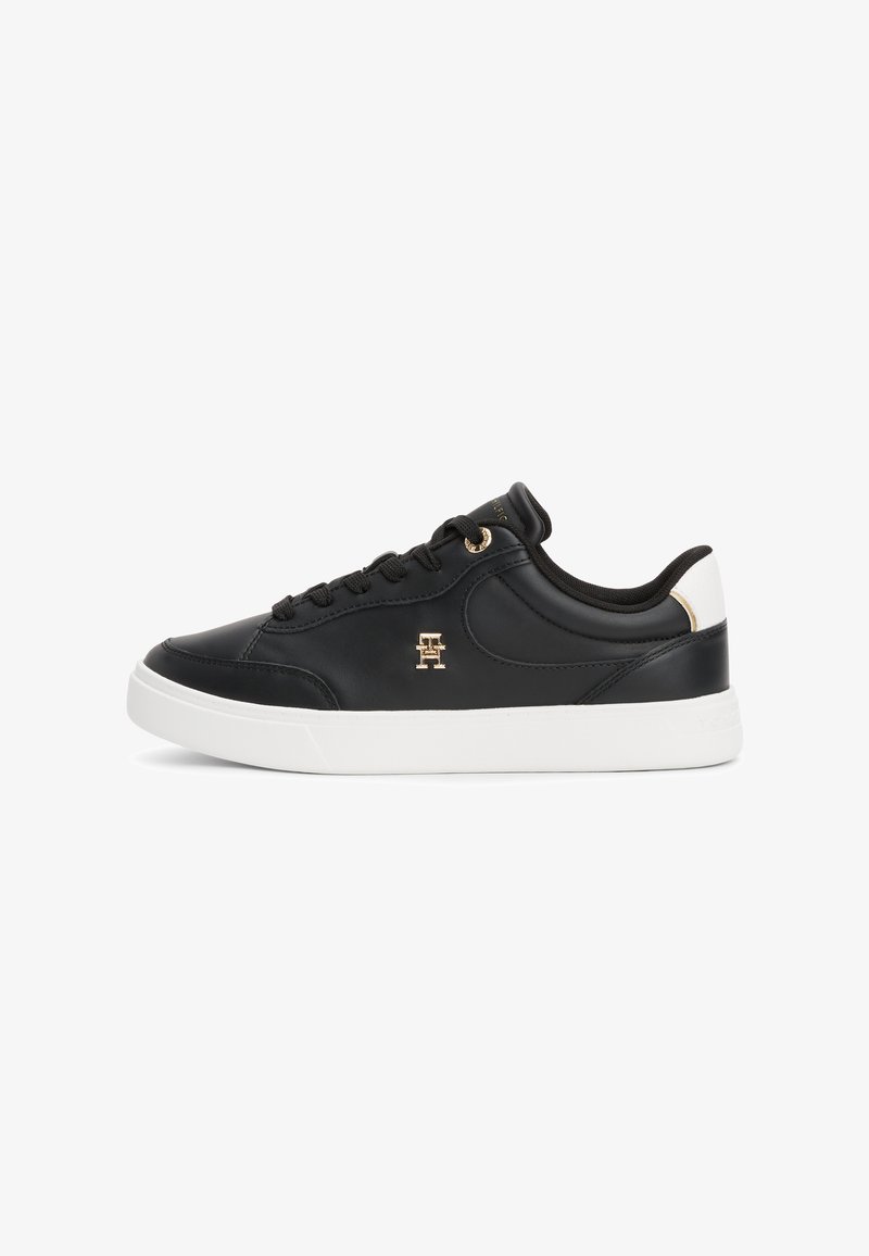 Black leather sneaker with white rubber sole, round toe, black laces, gold logo detail, and contrasting heel accent.