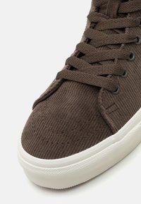 Sapatilhas de corduroy castanho com superfície texturizada, bico redondo e sola de borracha branca. Apresenta atacadores a condizer e ilhós de metal.