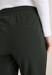 Donkergroene broek met een gestructureerde stof, elastische tailleband en twee achterzakken. Het ontwerp is gestroomlijnd met een subtiel patroon.