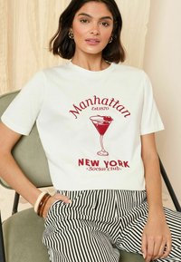 Biela tričko s grafickým dizajnom červenej martini poháre s nápisom "Manhattan Est. 1870" a textom "NEW YORK Social Club". Prúžkované čierno-biele nohavice.