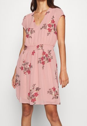 Femme portant une robe fleurie rose mi-longue aux manches courtes et à la taille froncée, debout devant un fond blanc uni.