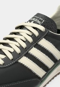 Black leather sneaker with white and gray accents, featuring three white stripes, cream laces, and embossed logo on the tongue.