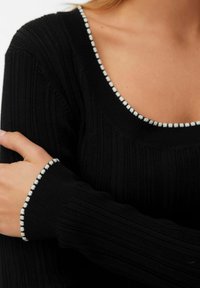 Svart ribbad stickad tröja med bred rundad halsringning och kontrasterande vit pom-pom kant vid halsen och ärmarna.