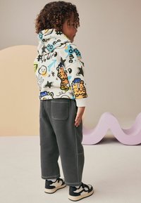 Barn iført en hvid hættebluse med farverige dyre- og doodle-tryk, parret med mørkegrå, løse bukser og sorte sneakers med hvide detaljer.