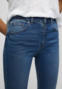 Vaqueros de denim azul de cintura alta con un ajuste slim, cinco bolsillos, costuras en contraste y una textura suave. Cierre de botón y cremallera en la parte delantera.