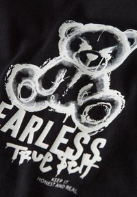 Σχέδιο αρκουδάκι σε λευκό στυλ σκίτσου πάνω από το κείμενο «FEARLESS True Self» και «KEEP IT HONEST AND REAL» σε μαύρο ύφασμα.