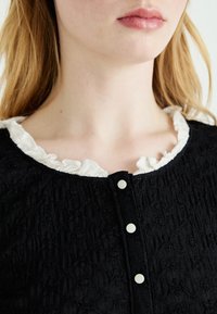 Černé texturované šaty s bílým volánkovým límcem; obsahují knoflíkovou légu se třemi bílými knoflíky na přední straně.