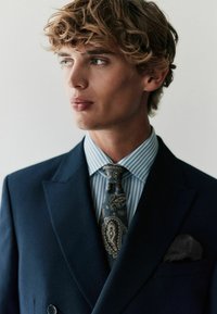 Tumesinine ülikond, millel on terav krae, koos helesinise ja valge triibulise särgi ning tumedast sinisest ja kuldsetest mustritest lipsuga.