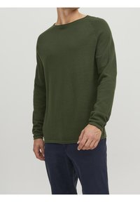 Grön långärmad tröja med en texturerad stickad design, rund halsringning och ribbade ärmslut, ihop med mörka byxor. Inga synliga logotyper eller mönster.