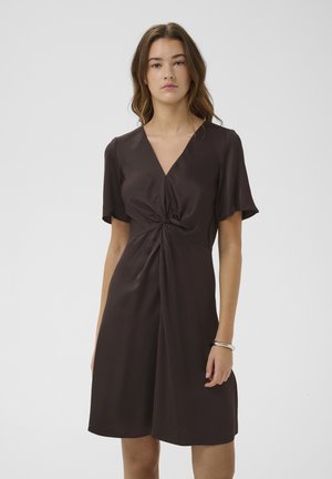 Noor naine kannab põlveni ulatuvat tumepruuni kleiti lühikeste varrukatega ja vöökohal keeratud sõlm-detailiga, seisab ühevärvilise tausta ees.