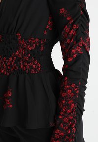 Haut noir à manches longues avec motifs floraux rouges, manches froncées et taille smockée créant une silhouette péplum ajustée.