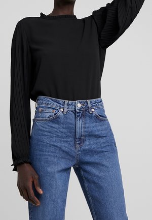 Person wearing a black long-sleeve pleated blouse tucked into high-waisted blue jeans with front pockets and belt loops.