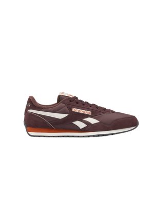 Zapatilla deportiva Reebok de color marrón oscuro con franjas blancas laterales, detalle naranja en la suela y cordones marrones, mostrada desde el lado izquierdo sobre un fondo blanco.