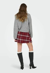 Mujer con cabello largo y ondulado, vestida con un suéter gris, falda de cuadros rojos, calcetines blancos y botas altas negras con tacón, de pie de espaldas.