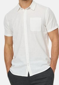 Mężczyzna w krótkim rękawie, biały koszula zapinana na guziki z kieszenią na piersi, ręce w kieszeniach ciemnoszarych spodni, na jednolitym tle.