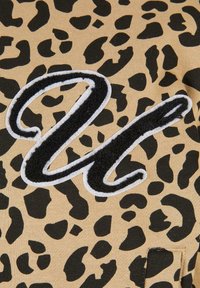 Tkanina s leopard uzorkom u bež i crnoj boji, s istaknutim crnim izvezenim slovom "U" obrubljenim belom bojom. Glatka tekstura, ležeran dizajn.