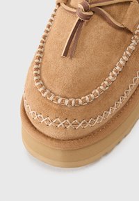 Tan suede moccasin with leather laces, decorative stitching, and a thick rubber sole. Features a rounded shape and textured surface.