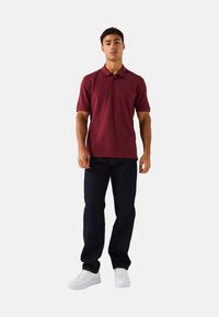 Camisa polo burdeos con cuello, mangas cortas y un branding sutil. Combinada con jeans oscuros y zapatillas blancas.