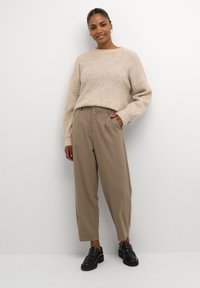 Béžový pletený svetr s dlouhými rukávy, v kombinaci s taupe skládacími kalhotami a černými mokasíny. Jednoduchý design, jemná textura a uvolněný střih.