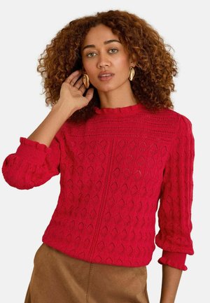 Roter gestrickter Pullover mit einem strukturierten Rautenmuster, geripptem Rundhalskragen und Rüsche an den Ärmeln. Das Model posiert mit der Hand nah am Gesicht.
