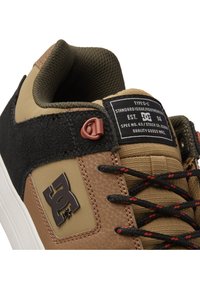 Brūni un zaļi sporta apavi ar teksturētu ādas virsu, melniem akcentiem un izteiktu logotipa uzlīmi. Ietver melnas auklas ar sarkaniem akcentiem.