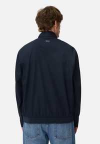 Mand med kort brunt hår iført en mørkeblå jakke og blå jeans, der står med ryggen til mod en ensfarvet baggrund.
