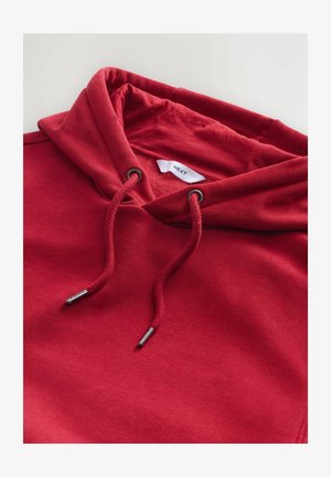 Raudona medvilninė džemperis su kapučinu, pasižymintis virvelėmis su sidabro spalvos metalinėmis detalėmis ir minkštu pamušalu. Etiketėje užrašyta "NEXT."