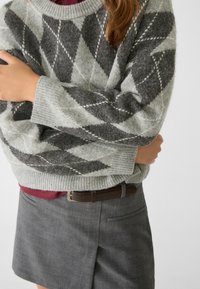 Helle graue Wolljacke mit einem Rautenmuster in Dunkelgrau und Weiß, gerippte Bündchen, getragen über einem weinroten Oberteil, kombiniert mit einem grauen Rock.