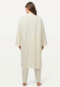 Lunga veste di colore crema, oversize con maniche larghe e taglio dritto, abbinata a pantaloni a sigaretta coordinati, caratterizzati da una texture morbida.