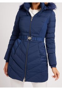 Mujer con un abrigo acolchado azul marino con capucha con pelo, cremallera frontal, cintura ceñida con cinturón y bolsillos laterales sobre una blusa blanca.