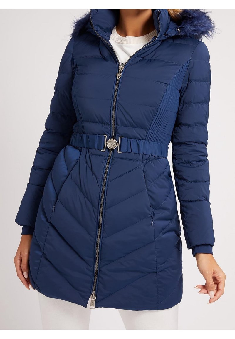 Mujer con un abrigo acolchado azul marino con capucha con pelo, cremallera frontal, cintura ceñida con cinturón y bolsillos laterales sobre una blusa blanca.