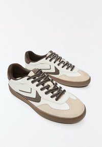 Paire de baskets basses beige, blanche et marron avec des lacets marron à motifs et des semelles en caoutchouc texturées sur fond blanc.