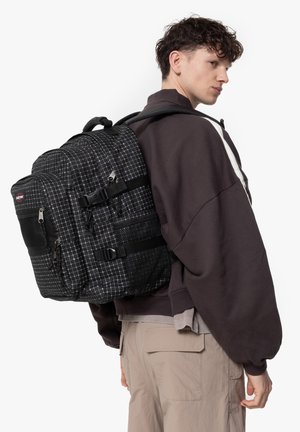 Black backpack with a grid pattern, multiple compartments, mesh accents, and adjustable straps, featured on a model wearing a brown sweater.