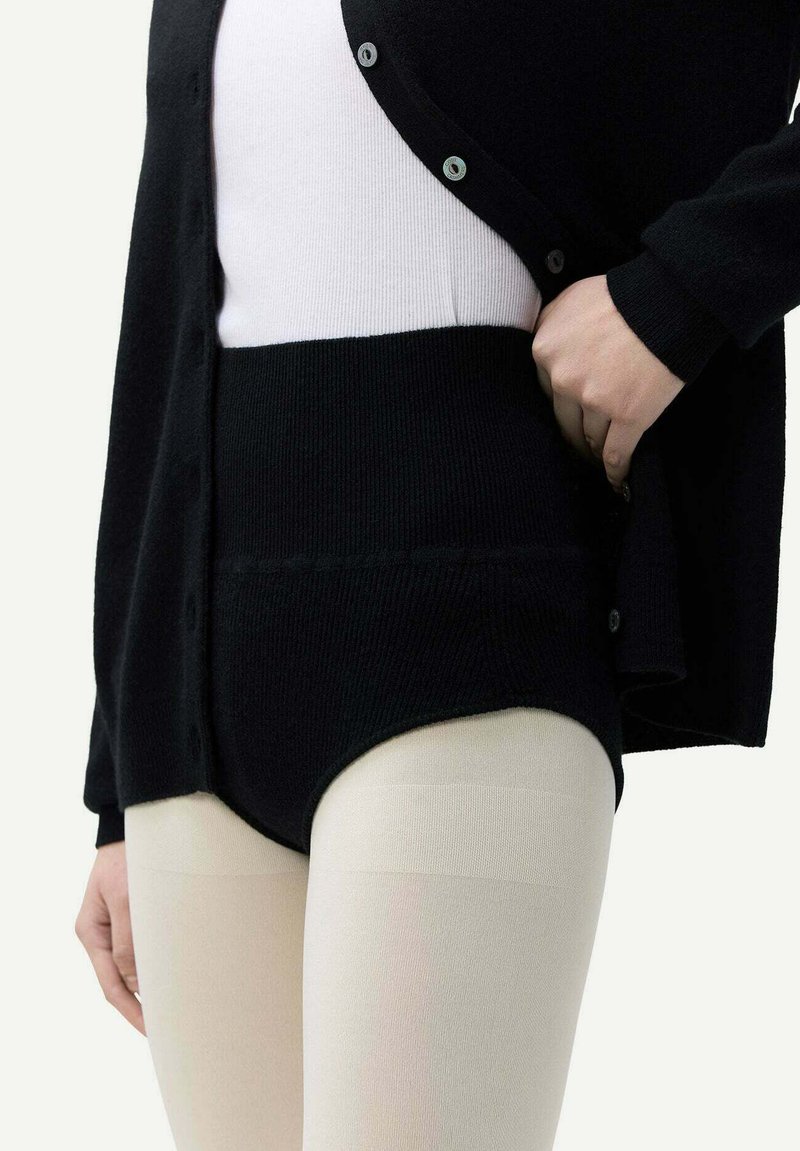 Svart, kraftig stickad kardigan med knappar, bärs över en vit topp och svarta högmidjade underkläder, matchat med ljusa strumpbyxor.