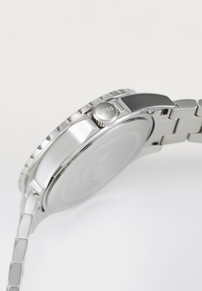 Montre en acier inoxydable argenté avec une lunette texturée, une couronne proéminente et un bracelet segmenté, présentant une finition polie et brossée.