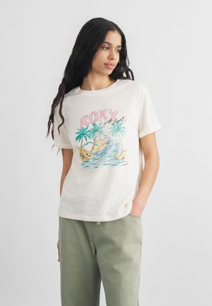 Kobieta z długimi ciemnymi włosami, ubrana w białą koszulkę z motywem tropikalnej plaży i napisem "ROXY z sercem", w połączeniu z jasnozielonymi spodniami.