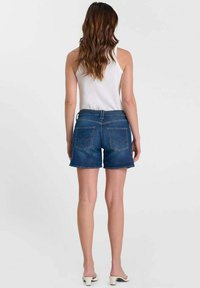 Femme aux longs cheveux bruns ondulés portant un haut blanc sans manches, un short en jean bleu et des sandales blanches à talons, vue de dos sur un fond uni.