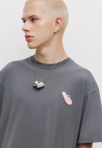 Grå oversized T-shirt med broderi af en pink og blå flip-flop på brystet. En kraftig sølvhalskæde bæres om halsen.