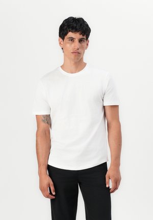 T-shirt en coton blanc avec un col rond, des manches courtes et une coupe décontractée. Porté avec un pantalon noir, présentant une texture lisse subtile.
