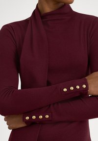 Μπορντό μακρυμάνικη μπλούζα με ψηλό λαιμό, ριμπ ύφανση και χρυσά κουμπιά στα μανίκια. Διαθέτει λεπτή, εφαρμοστή γραμμή.