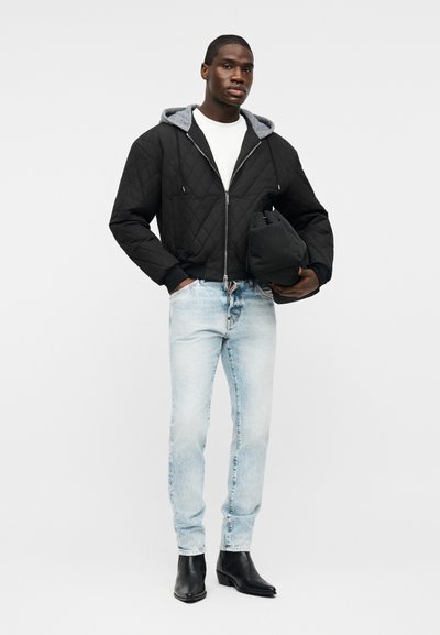 Homme portant une veste matelassée noire avec capuche grise, chemise blanche, jean bleu clair, bottes noires, tenant un sac noir sur fond blanc.