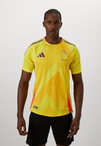Gul sportstrøje med diagonale mønstre i mørkere gult og lilla, med røde accenter, korte ærmer og Adidas-logo på brystet.