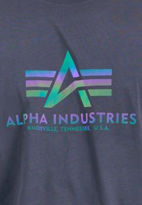 Mørkegrå T-shirt med et flerfarvet holografisk logo med teksten "ALPHA INDUSTRIES" og "Knoxville, Tennessee, U.S.A." nedenunder.