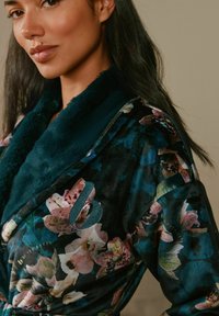 Woman with long dark hair wearing a dark floral patterned robe with teal fur collar, shown from the shoulders up, slight smile.