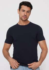 Mężczyzna o ciemnych włosach, ubrany w jednolity granatowy t-shirt i jasnoniebieskie dżinsy, poprawiający hem t-shirtu obiema rękami.