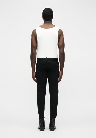 Homme debout de dos portant un haut blanc sans manches, un pantalon noir et des bottines noires sur un fond blanc uni.